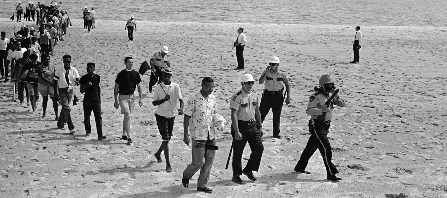 Black & white photo of police parading people in a line from the beach